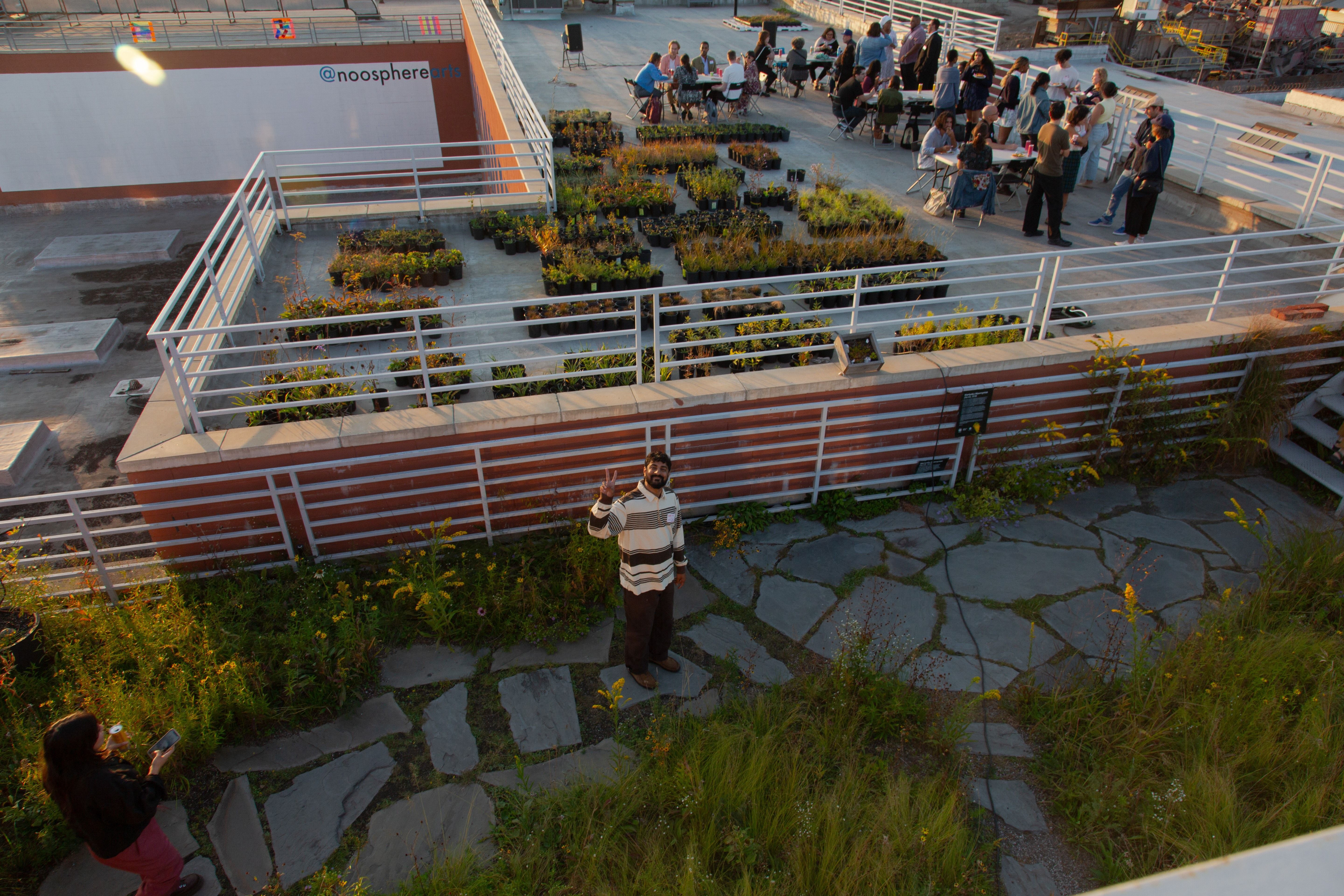 A man giving a peace sign in the foreground, while a party happens on a lower level in the background.