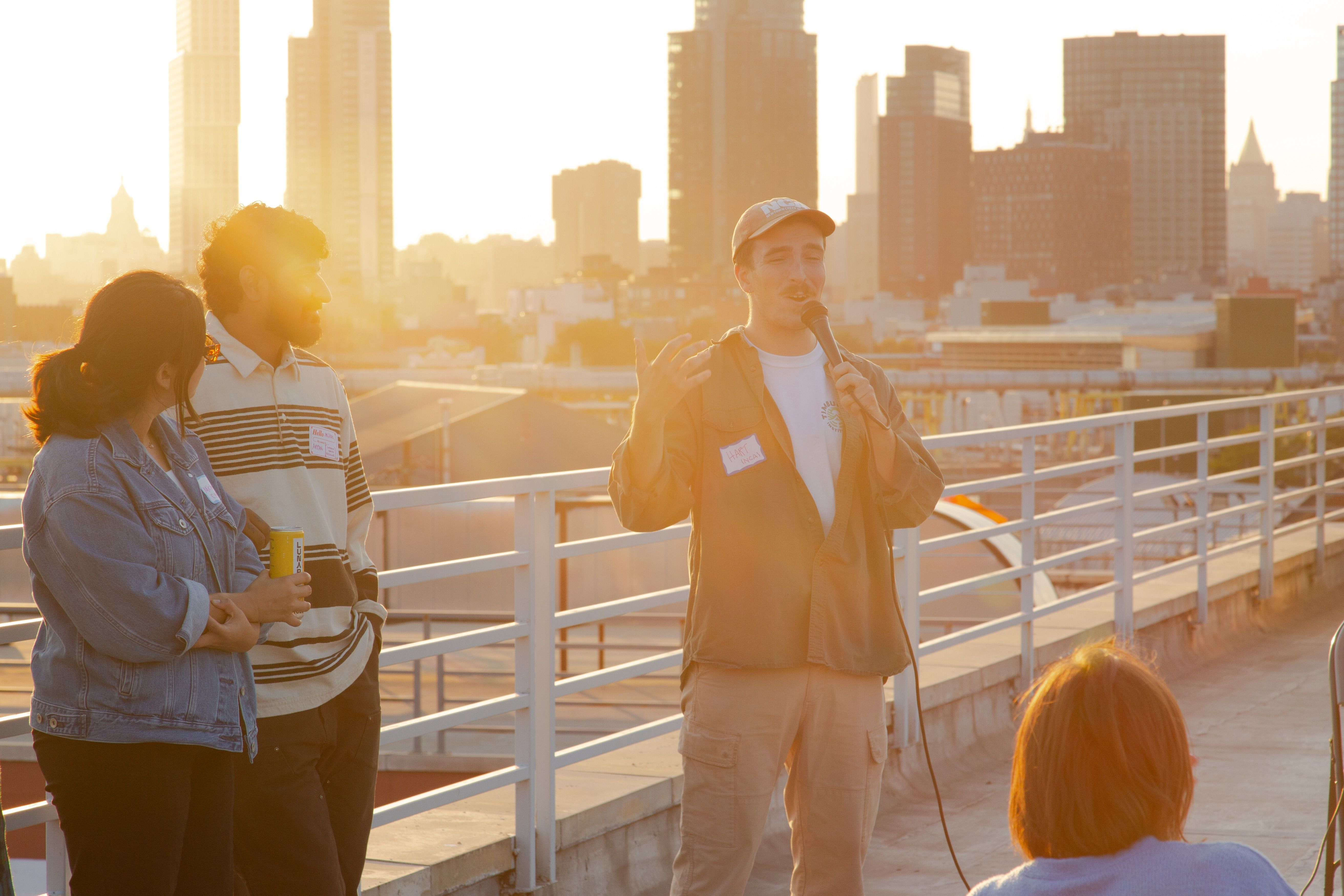 Hart Mankin speaking on a microphone with sunset-lit city skyline behind them.