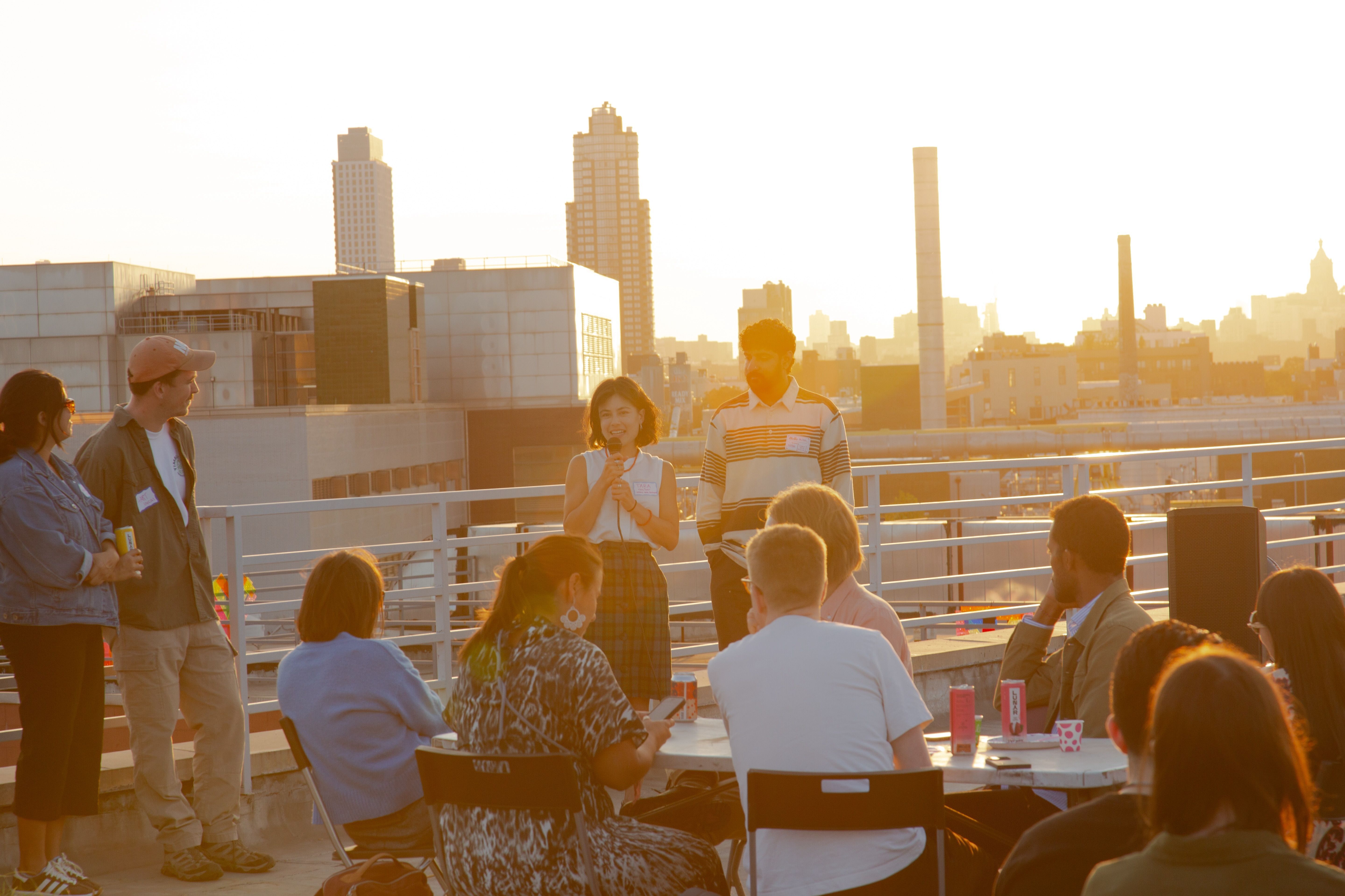 Tara Pham speaking on a microphone, alongside Arpan Somani, as they address an audience with sunset-lit city skyline behind them.